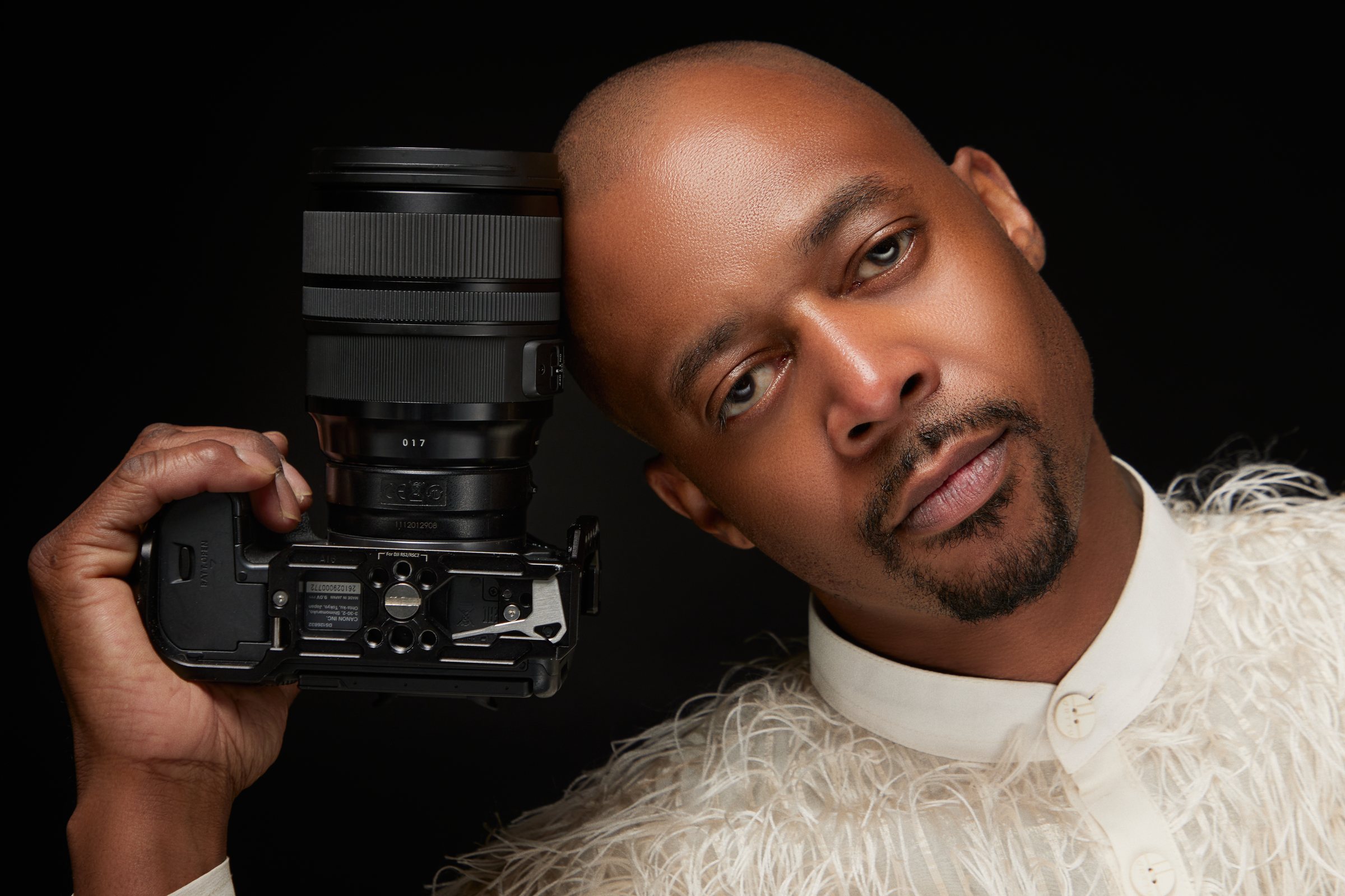 Skyye Kimmons holding a Canon camera against the side of his face, wearing a cream feathered shirt against a black background